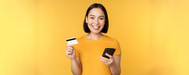 Zakupy online wesoła Azjatka trzymająca kartę kredytową i smartfona płacącego zamówienie za pomocą telefonu komórkowego