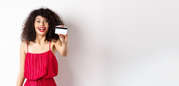 Bezpłatne zdjęcie zakupy podekscytowane stylową damą w czerwonej sukience z makijażem, pokazującą plastikową kartę kredytową i uśmiechniętą zdumioną s