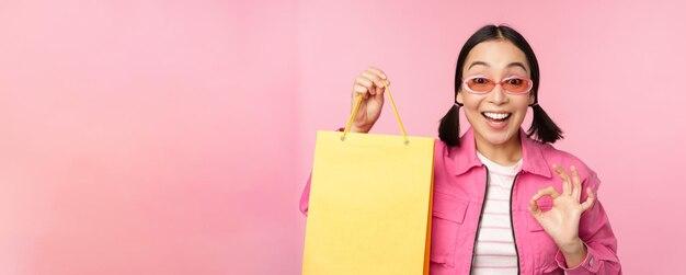 Zakupy Stylowa Azjatycka dziewczyna w okularach przeciwsłonecznych pokazująca torbę ze sklepu i uśmiechnięta polecająca promocję sprzedaży w sklepie stojącą na różowym tle
