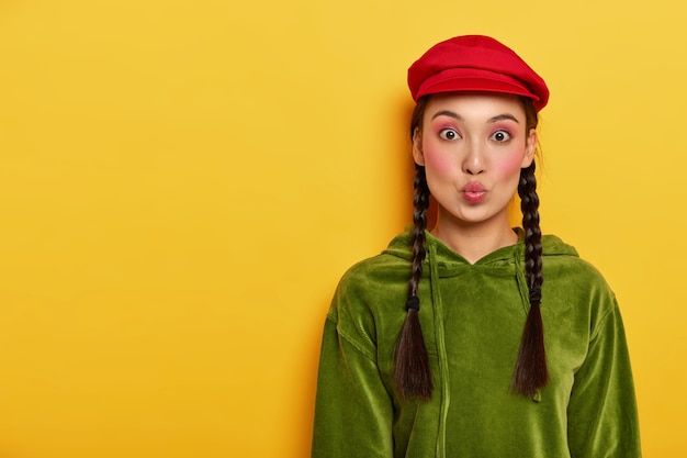 Zalotna piękna nastolatka marszczy usta, nosi jasny makijaż, ubrana w stylową czerwoną czapkę i sztruksową bluzę, dwa warkocze