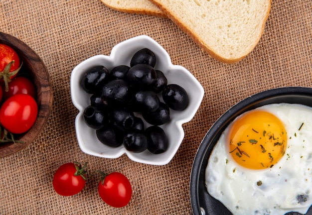 Zamknąć widok czarnych oliwek w misce z pomidorami, chlebem i jajkiem sadzonym na worze