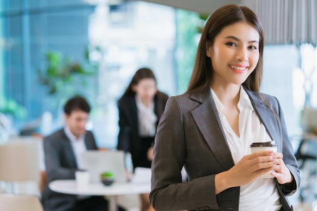 Zamknij się inteligentny atrakcyjny azjatycki biznes kobieta uśmiech ręka trzyma gorący napój z nowoczesnym tłem biurowym
