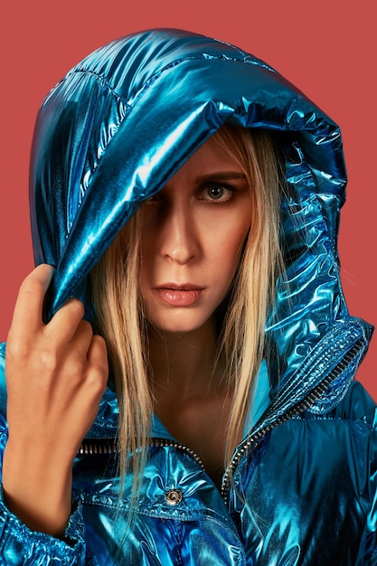 Zamknij się moda zdjęcie młodej kobiety, zakrywając się kapturem. Nosi niebieską puchową kurtkę oversize w studio na czerwonym tle