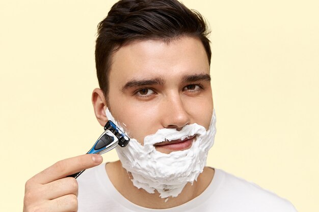 Zamknij się obraz przystojnego młodego ciemnowłosego mężczyzny z białą pianą na twarzy, do golenia z jednorazową brzytwą do golenia z ziarnem.