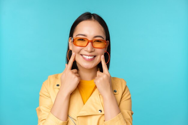 Zamknij się portret azjatyckiej młodej kobiety w okularach przeciwsłonecznych uśmiechniętej i wyglądającej romantycznie stojącej szczęśliwej na niebieskim tle
