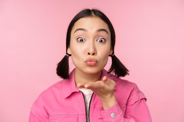 Zamknij się portret pięknej głupiej azjatyckiej dziewczyny wysyłającej powietrze pocałunek mwah w kamerę, która wieje stojąc na różowym tle