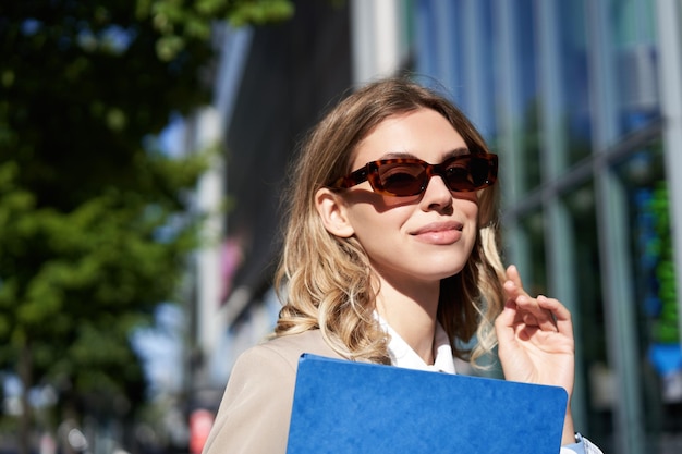 Bezpłatne zdjęcie zamknij się portret uśmiechniętej młodej kobiety w okularach przeciwsłonecznych w firmowym garniturze, trzymając folder z dokumentem roboczym