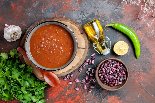 Zamknij widok klasycznej zupy pomidorowej w niebieskiej misce łyżka na drewnianej tacy butelka oleju czosnek sól i cytryna pęczek zieleni na stole mieszanym