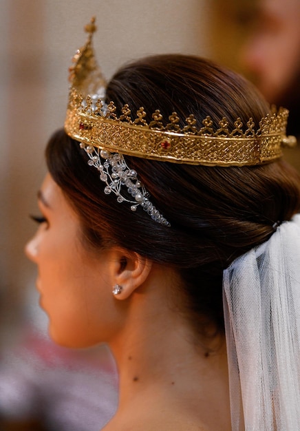 Bezpłatne zdjęcie zamknij widok ładnej kobiety ze stylową fryzurą i naturalnym makijażem, ubranej w srebrne dodatki na uszach, trzymającej złotą koronę na głowie podczas ceremonii ślubnej w kościele