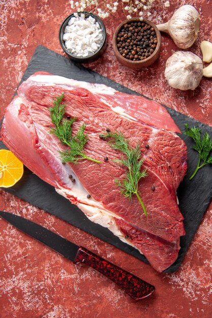 Zamknij widok świeżego czerwonego mięsa z zielonym i pieprzem na czarnej desce nóż czosnek cytryna przyprawy drewniany młotek cytryna na pastelowym czerwonym tle oleju