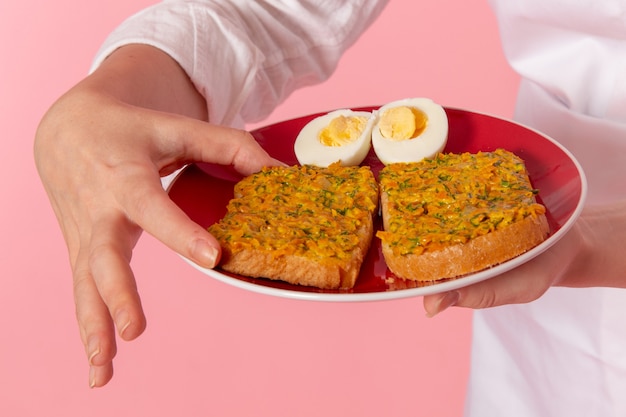 Zamknij widok z przodu kobieta cukiernik w białym zużyciu trzymając talerz z tostami i jajami na różowej ścianie praca kucharz kuchnia kuchnia jedzenie
