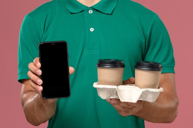 Zamknij widok z przodu kurier męski w zielonym mundurze, trzymając brązowe filiżanki kawy dostawy i telefon na różowym biurku
