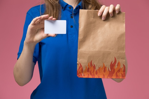 Zamknij widok z przodu żeński kurier w niebieskim mundurze, trzymając białą kartę i pakiet żywności na różowym świetle biurka jednolita praca firmy
