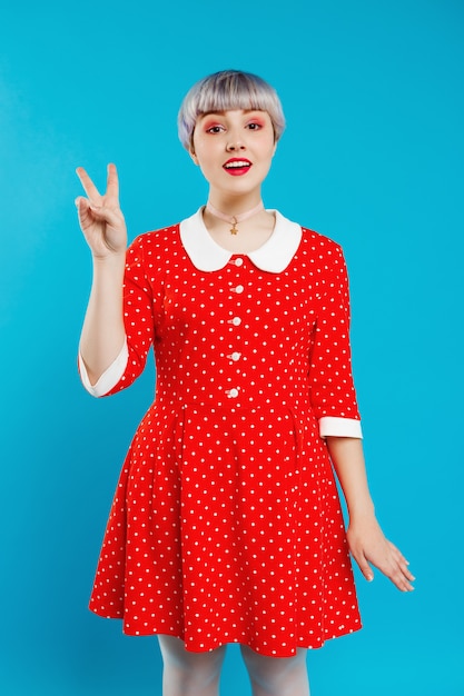 Zamyka w górę portret pięknej lalkowatej dziewczyny z krótkim jasnofioletowym włosy jest ubranym czerwieni suknię pokazuje zwycięstwo gest nad błękit ścianą