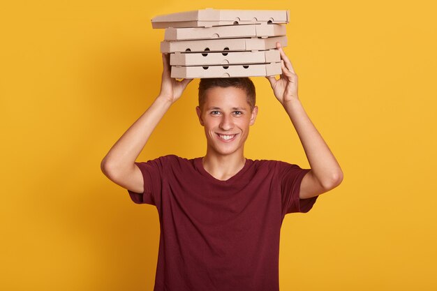 Zamyka w górę portreta młody rozochocony doręczeniowy mężczyzna z czerwoną koszulki pozycją, trzyma stertę kartonowi pudełek po pizzy na szarości ścianie.