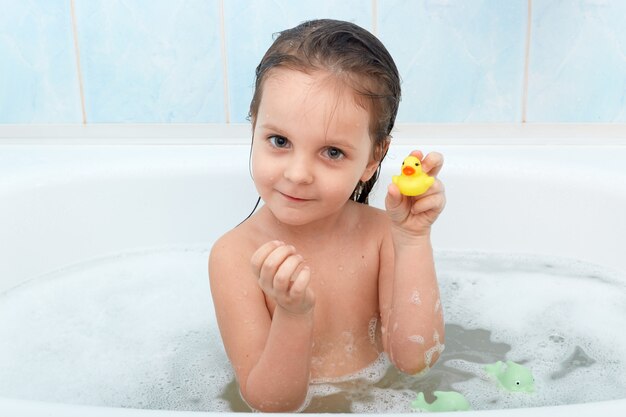 Zamyka w górę portreta szczęśliwy powabny małej dziewczynki obsiadanie w kąpielowej balii bawić się z żółtą kaczką w łazience.