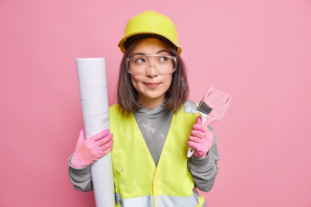 Zamyślona azjatycka konserwatorka trzyma zrolowany pędzel do malowania planów odwraca wzrok w zamyśleniu nosi jednolite pozy na różowo