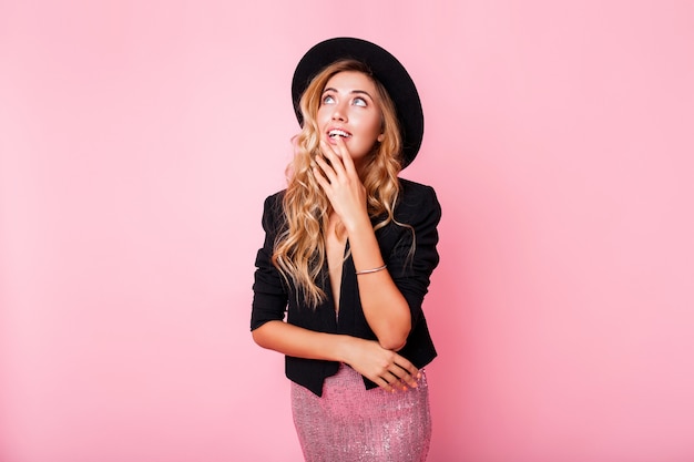 Bezpłatne zdjęcie zamyślona blondynka w czarnym kapeluszu pozowanie na różowej ścianie. ubrana w modną sukienkę z sekwencją, czarną marynarkę i czapkę.