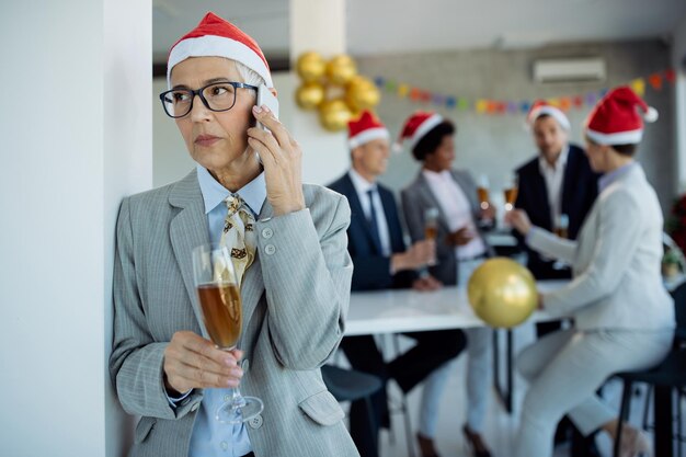 Zamyślona dojrzała bizneswoman rozmawia przez telefon będąc na przyjęciu bożonarodzeniowym w biurze