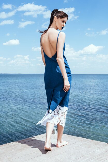 Zamyślona dziewczyna w długiej dżinsowej sukience z tyłu boso. Kobieta na molo nad morzem w lecie. Model z kucykiem