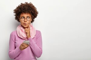 Bezpłatne zdjęcie zamyślona kobieta patrzy na bok, trzyma telefon komórkowy, czeka na telefon, zaciska usta
