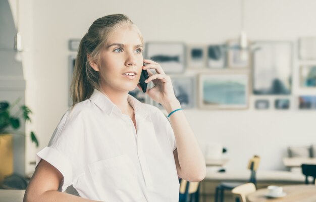 Zamyślona piękna młoda kobieta ubrana w białą koszulę, rozmawiając przez telefon komórkowy, stojąc w przestrzeni coworkingowej i odwracając wzrok