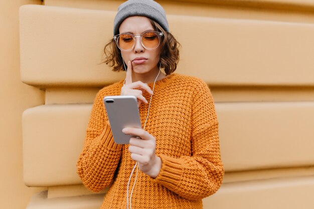 Zamyślona śliczna kobieta w dużym swetrze patrząc na ekran telefonu idąc ulicą