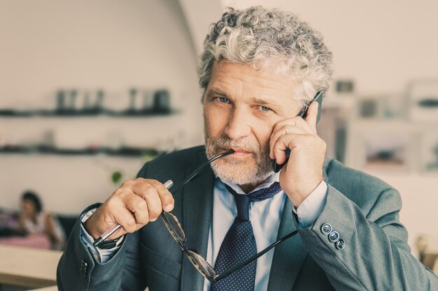 Zamyślony dojrzały biznesmen rozmawia przez telefon komórkowy, stojąc przy co-working, opierając się na biurku