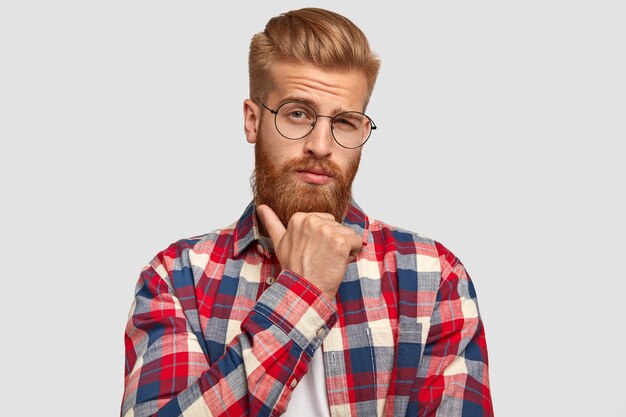 Bezpłatne zdjęcie zamyślony nieogolony mężczyzna trzyma podbródek, patrzy w zamyśleniu prosto w kamerę, myśli o czymś ważnym, ubrany w stylową kraciastą koszulę