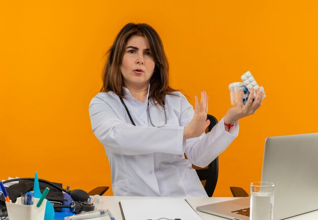 Zaniepokojona kobieta w średnim wieku ubrana w szlafrok medyczny ze stetoskopem siedząca przy biurku na laptopie z narzędziami medycznymi trzymającymi pigułki i pokazująca gest stop na odizolowanej pomarańczowej ścianie