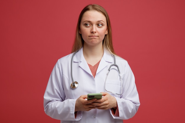 Zaniepokojona młoda blond lekarka ubrana w szlafrok medyczny i stetoskop na szyi trzymająca telefon komórkowy obiema rękami patrząc w bok z zaciśniętymi ustami