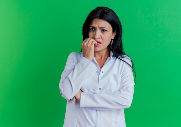Zaniepokojona młoda kobieta lekarz ubrana w szlafrok medyczny patrząc na bok kładąc palce na wardze na białym tle na zielonej ścianie z miejsca na kopię