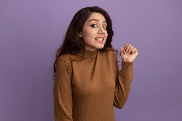 Zaniepokojona młoda piękna dziewczyna ubrana w brązowy sweter z golfem wskazuje z tyłu na białym tle na fioletowej ścianie z miejsca na kopię