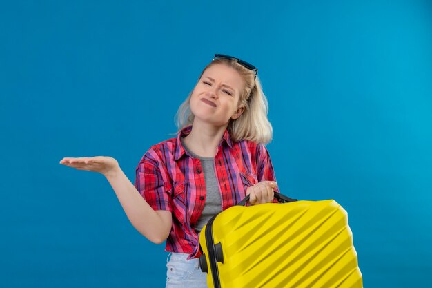 Zaniepokojona młoda podróżniczka ubrana w czerwoną koszulę i okulary na głowie trzymająca walizkę wskazuje na bok na odizolowanej niebieskiej ścianie