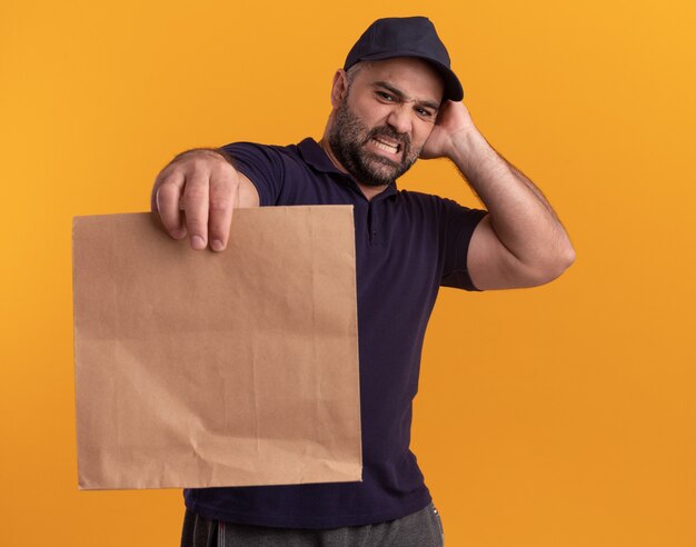Zaniepokojony mężczyzna w średnim wieku w mundurze i czapce trzymający papierowe opakowanie żywności i patrząc na niego, kładąc dłoń na głowie na białym tle na żółtej ścianie