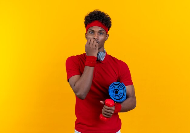 Zaniepokojony młody afroamerykański sportowy mężczyzna w opasce na głowę i opasce ze słuchawkami trzymającymi matę do jogi i gryzie paznokcie na białym tle na żółtym tle