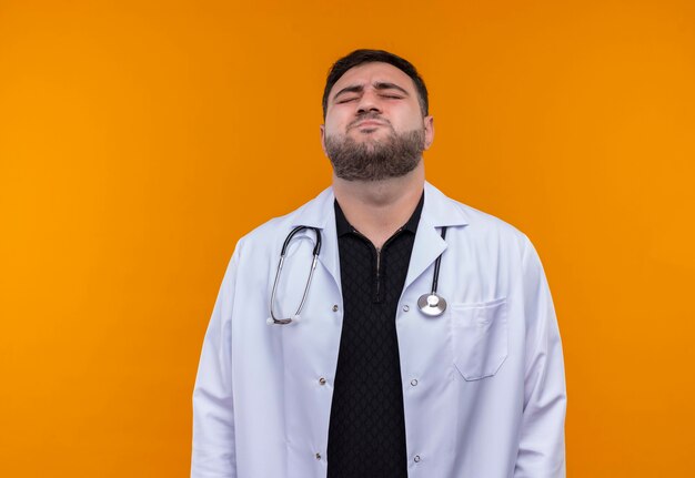 Zaniepokojony młody, brodaty lekarz w białym fartuchu ze stetoskopem wyglądający na zmęczonego i zestresowanego zamykającego oczy