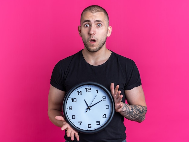 Zaniepokojony młody przystojny facet ubrany w czarną koszulkę trzymającą zegar ścienny na białym tle na różowym tle