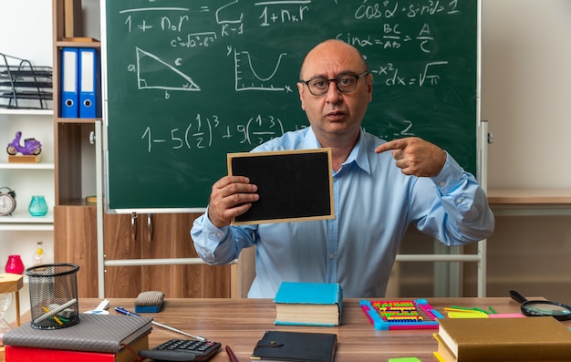 Zaniepokojony nauczyciel w średnim wieku siedzi przy stole z przyborami szkolnymi i wskazuje na mini tablicę w klasie