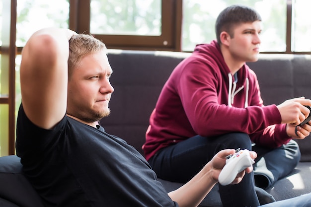 Zapaleni gracze. Dwóch młodych szczęśliwych mężczyzn grających w gry wideo siedząc na kanapie
