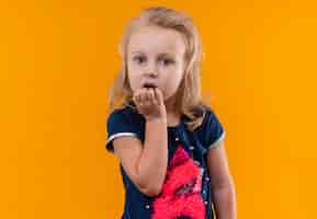 Bezpłatne zdjęcie zaskakująca piękna mała dziewczynka o blond włosach w granatowej koszuli trzymająca rękę na brodzie na pomarańczowej ścianie