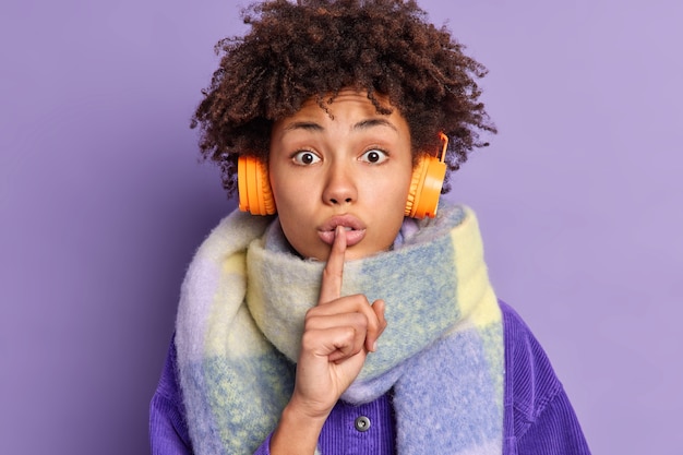 Zaskoczona Afroamerykanka Przykłada Palec Wskazujący Do Ust I Robi Gest Ciszy, Mówi Sekret, Nosi Ciepły Szalik Na Szyi, Słucha Muzyki Przez Bezprzewodowe Słuchawki.