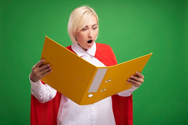 Zaskoczona Blondynka Superbohaterka W średnim Wieku W Czerwonej Pelerynie, Trzymająca I Patrząca Na Folder Odizolowany Na Zielonej ścianie