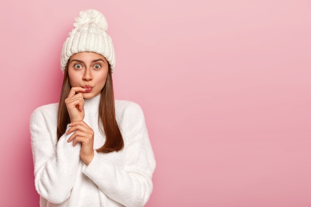 Zaskoczona ciemnowłosa kobieta wygląda zaskakująco, ma zaokrąglone usta, nosi śnieżnobiałą zimową czapkę i sweter, ubrana w ciepły strój pozuje na różowej ścianie