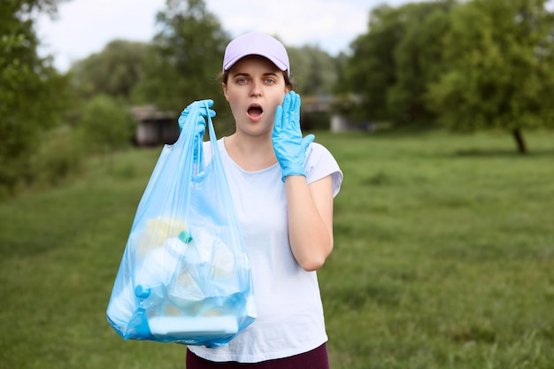 Zaskoczona dziewczyna z szeroko otwartymi ustami stoi z workiem na śmieci w dłoni, trzymając dłoń na policzku