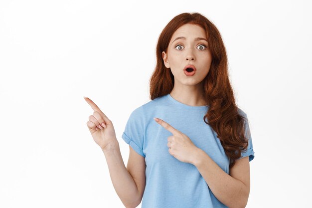 Zaskoczona i podekscytowana rudowłosa kobieta opowiadająca ważne wiadomości, wskazując palcami na bok w lewym górnym rogu baner z logo, dysząca i mówiąca „wow”, pod wrażeniem reklamy, białe tło