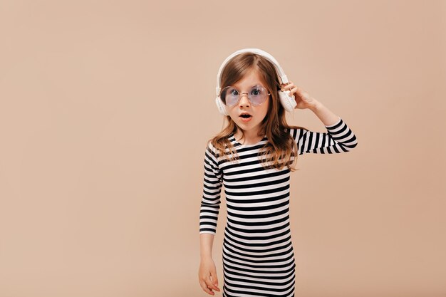 Zaskoczona mała stylowa dziewczyna ubrana w sukienkę w paski i modne okrągłe szkło, słuchając muzyki i pozując do kamery