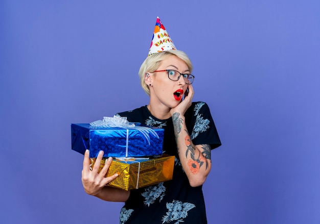 Zaskoczona młoda blondynka imprezowa w okularach i czapce urodzinowej trzymająca pudełka z prezentami, patrząc na kamerę, trzymając rękę na twarzy odizolowaną na fioletowym tle z miejsca na kopię