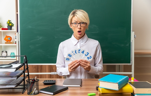 Bezpłatne zdjęcie zaskoczona młoda blondynka nauczycielka w okularach siedząca przy biurku z szkolnymi narzędziami w klasie pokazująca fanów liczb