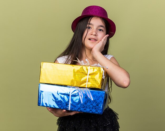 zaskoczona młoda dziewczyna kaukaska z fioletowym kapeluszem strony, kładąc rękę na twarzy i trzymając pudełka na prezenty odizolowane na oliwkowej ścianie z miejsca na kopię